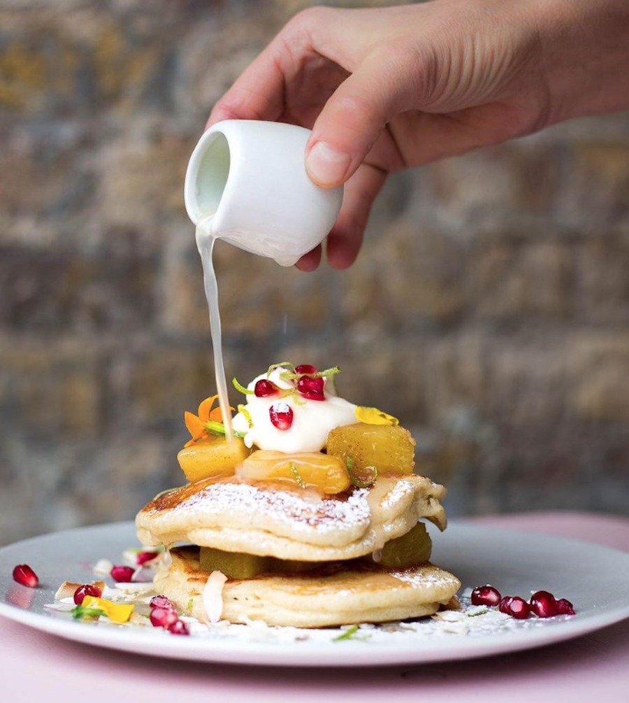 This is an image of cute little round pancakes on a white plate with pomegranate seeds on top. A hand is pouring out a syrup liquid on top of the pancakes. There is a brick wall in the background that is out of focus and the table under the plate is pink 