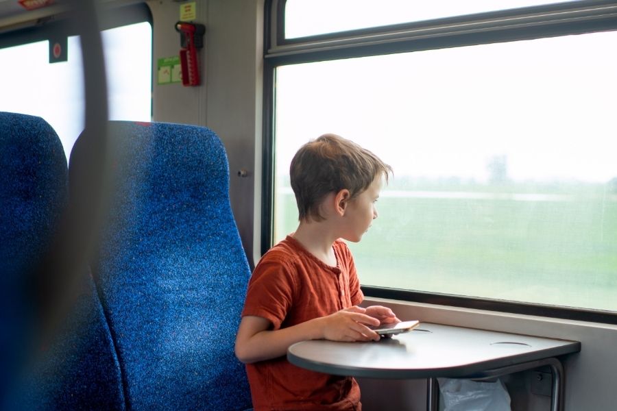 A young boy in a rust-coloured t-shirt sits alone at a train table seat, looking out the window at a green countryside landscape passing by.