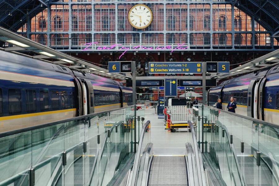 The interior of St Pancras International station showing two Eurostar trains on either side of platforms 5 and 6, with the station's ornate Victorian brick facade, large clock, and a pink neon art installation visible in the background.