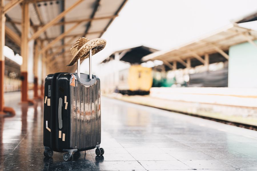 A black hard-shell suitcase with stickers and a straw hat resting on its handle, standing on a station platform with a blurred yellow and green train in the background.