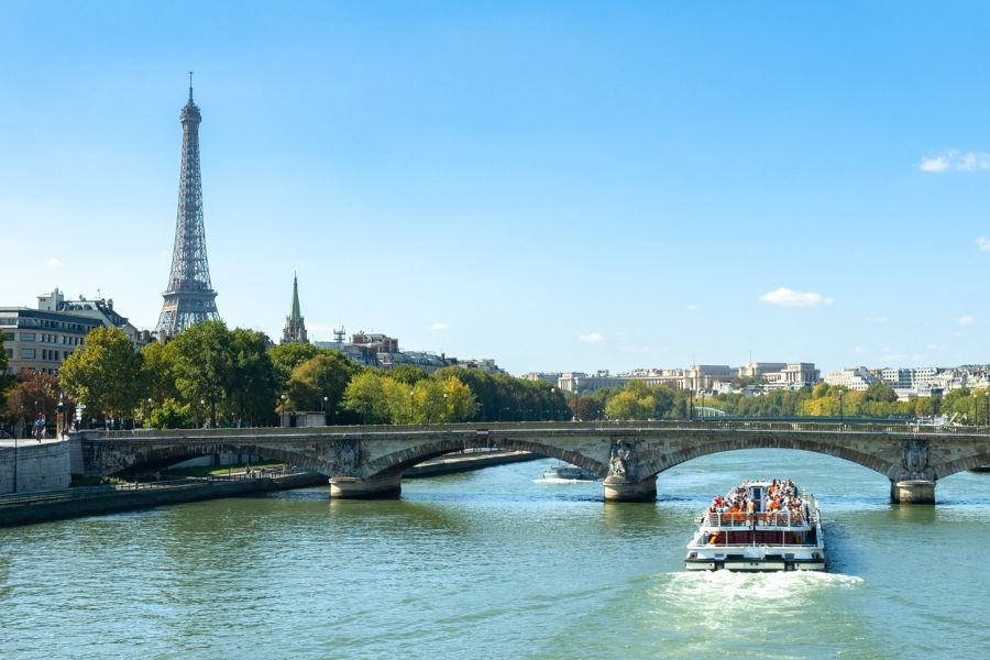 A river cruise boat travels along the Seine in Paris on a sunny day, with the Eiffel Tower and the city's rooftops visible beyond the trees and a stone bridge in the foreground.