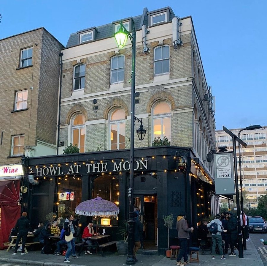 This is an image of a trendy pub in London. It is a low square shaped building painting black. Behind the pub is a tall building that looks like apartment blocks. The sky is bright blue and it appears to be nearing to dusk. 