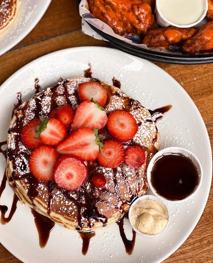 This is an image of pancakes on a white plate. On top of the pancakes are sliced strawberries topped with powdered sugar and chocolate sauce. The table underneath is wooden. 