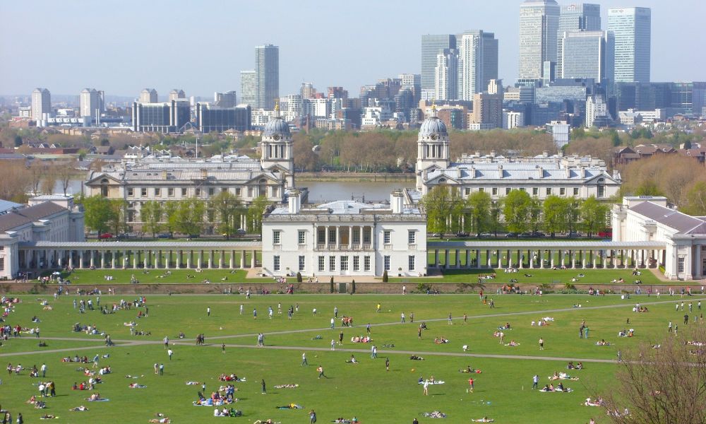 This is an image of a big park with a view of a house down the bottom and sprawling green gardens with people lounging and picnicing.