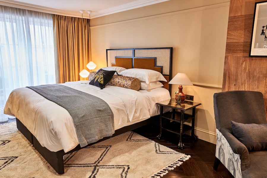 This is an image of a beautiful hotel bedroom with a big double bed that is neatly made, warm lighting, a neutral palette and quite minimalistic and sleek furniture. It is cozy and warm and neutral in aesthetic.