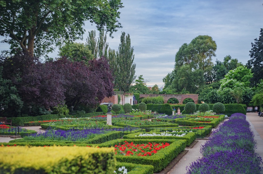 This is an image of immaculately kept gardens in a park. There are pretty flower beds in different colours nicely lined up next to one another and a soft blue sky in the background.