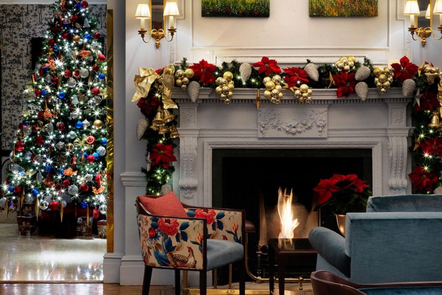 This is a picture of a lit up fire place with baubles and Christmas decorations surrounding. Behind it is a Christmas tree shining brightly.