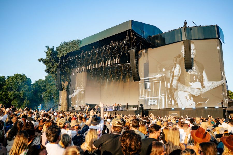 Large outdoor concert stage at BST Hyde Park with a guitarist projected on the giant screen, surrounded by a crowd in summer sunshine.