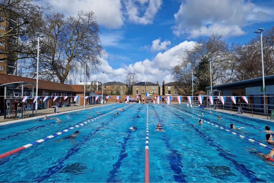 Outdoor swimming lido in London with swimmers in blue-tiled lanes under a bright cloudy sky, surrounded by trees and red-brick buildings.