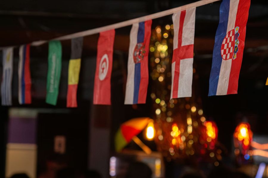 String of international football team flags as bunting hanging inside a dimly lit pub, with warm lights in the background.