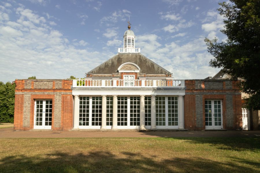 The Serpentine North Gallery building in Kensington Gardens on a sunny day, a red-brick neoclassical pavilion with white columns and a domed cupola.