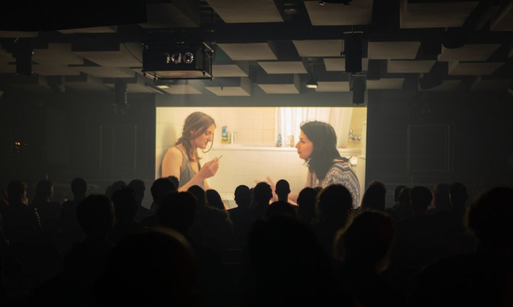 This is an image of a full cinema with an indie film playing in front of them with two women talking to one another.