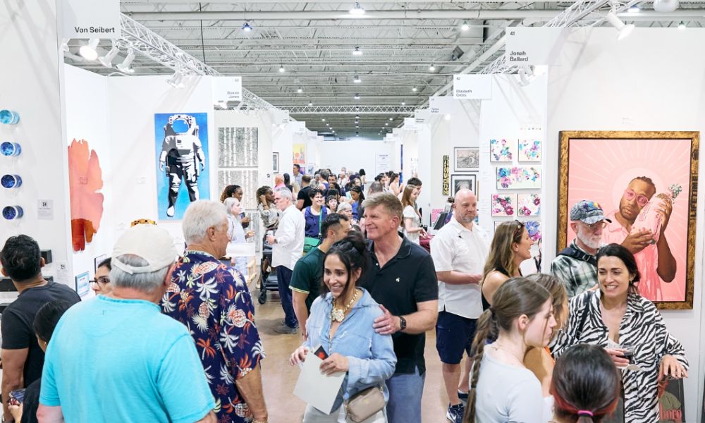 This is an image of an art fair with people walking around looking at pieces hung up on walls.