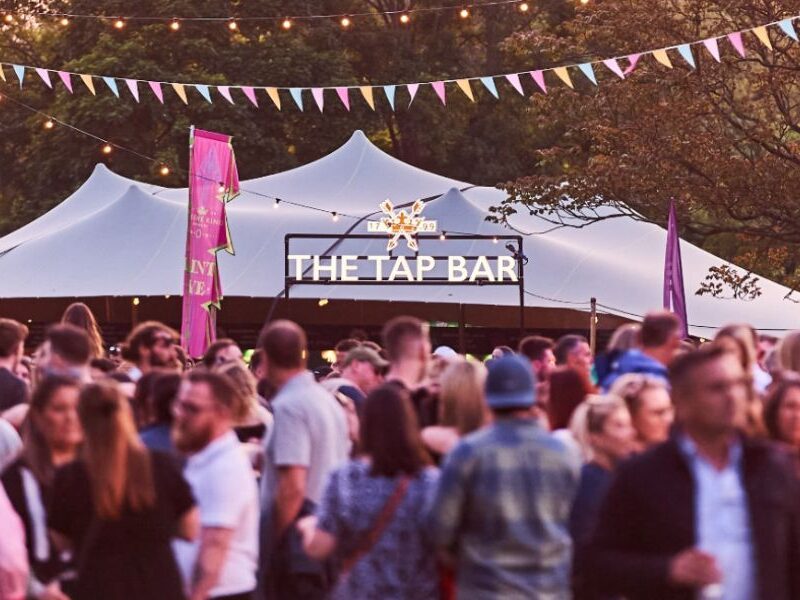 This is an image of a group full of people all partying and dancing in front of a big tap bar tent where they are serving drinks.