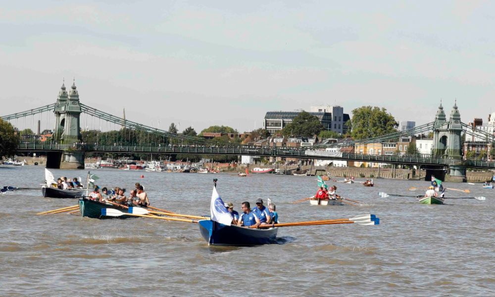 This is an image of people on boats riding down the Thames with ores. There is a bridge in the background.