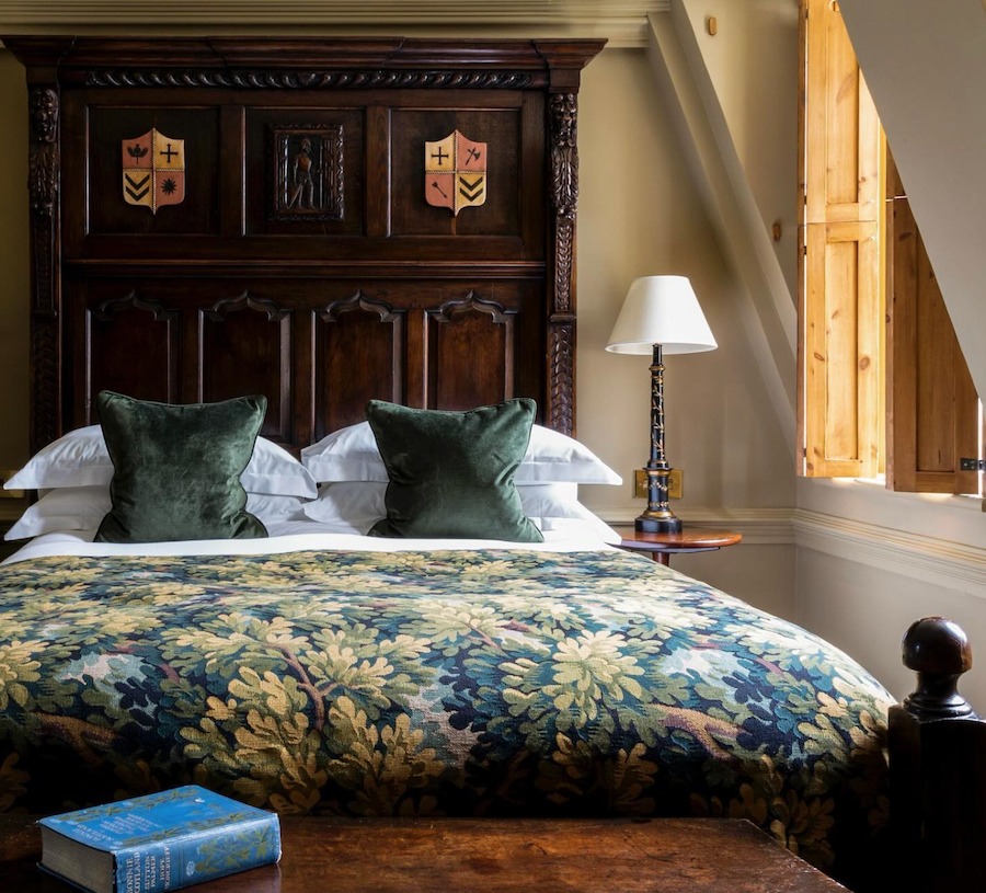 This is an image of a decadent double bed with green and blue bedding of a tree pattern and a wooden headboard behind the bed.