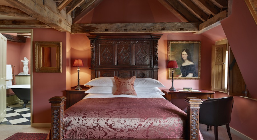 This is an image of a decadent looking boutique hotel room with a double bed in the middle of the room that has a dark mahogany headboard. The bedding is mainly white with a dark pink, velvet pillow and throw on top of the bed. There is an old fashioned portrait handing on the wall by the side of the bed. The walls are painted in a similar colour to the pink pillow on the bed and there are wooden beams on the ceiling giving the room a very old fashioned aesthetic and feel. We can also see into the bathroom that has black and white tiling and a white marble sculpture by the Victorian looking bath.