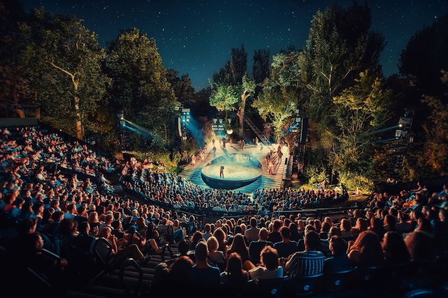 A semi-circular stage with bright lights in the center of a park theatre. There are actors performing on the stage in front of a large audience.