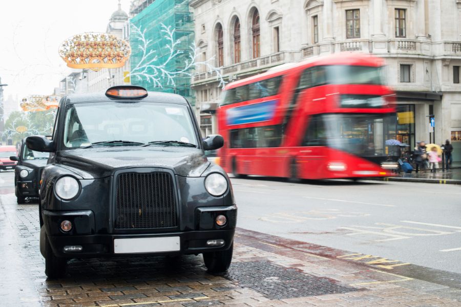 A black London taxi cab parked on a wet city street with a red double-decker bus passing in the background