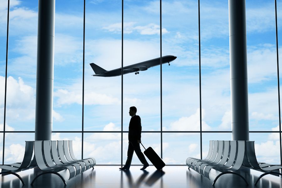 A silhouette of a traveller walking through a bright airport terminal with a suitcase, with a plane visible taking off through the floor-to-ceiling windows
