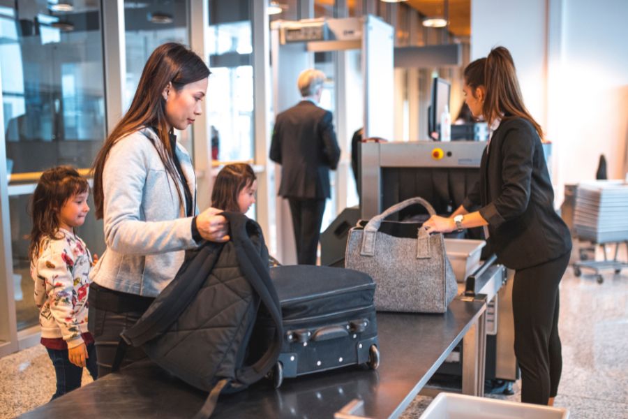 A woman and two young children placing bags onto a security conveyor belt at airport security, assisted by a member of staff