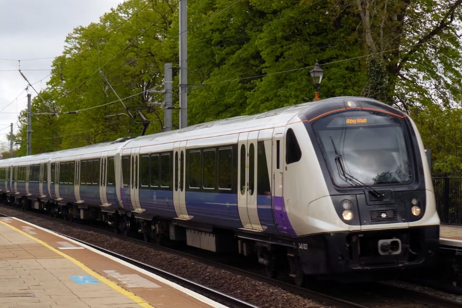 An Elizabeth line train in its distinctive white and purple livery pulling into a station, surrounded by green trees