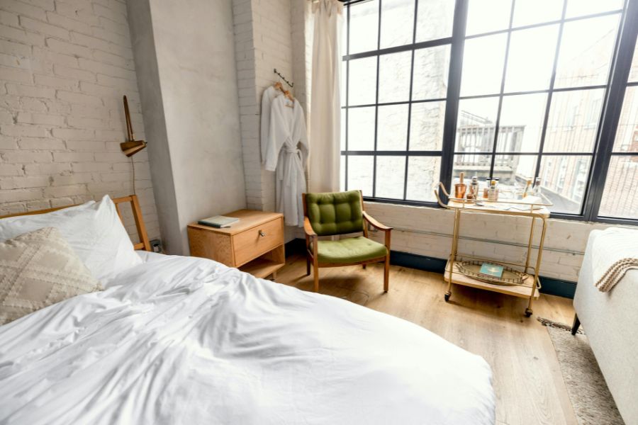 A stylish boutique hotel room with exposed white brick walls, a rumpled white bed, a retro green armchair, a gold bar cart, and large industrial-style windows.