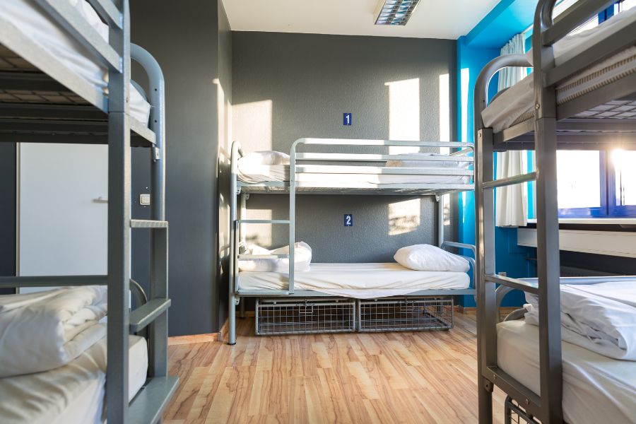 A hostel dormitory with rows of metal bunk beds, numbered berths, wire storage baskets underneath, and blue and grey walls with large windows.