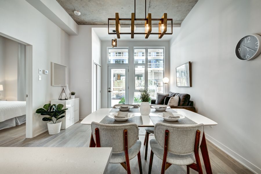 A bright, contemporary open-plan apartment with a white dining table set for four, a statement geometric pendant light, and large glass doors leading to an outdoor walkway.