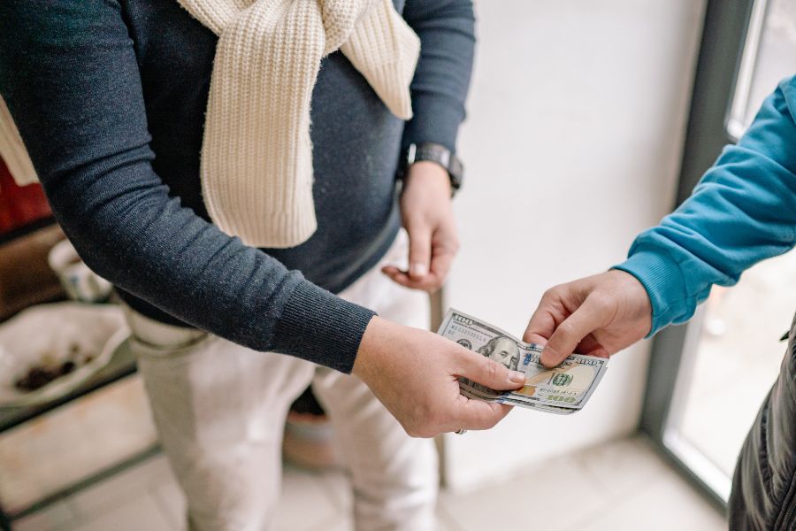 A close-up of two people exchanging US dollar banknotes, with one person wearing a cream scarf and the other in a blue jumper.