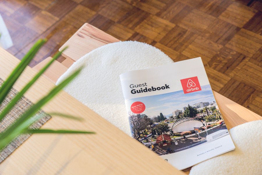 An Airbnb Guest Guidebook resting on a wooden coffee table beside a white fluffy rug and a green plant, with a parquet floor in the background.