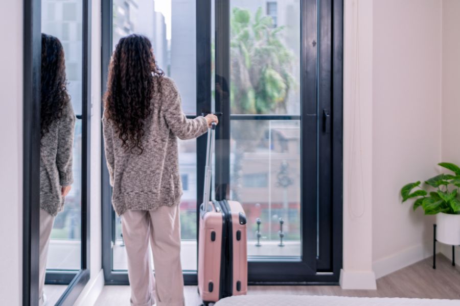 A woman with long curly hair standing at a floor-to-ceiling glass door with a pink suitcase, looking out at a city view from inside a modern apartment.