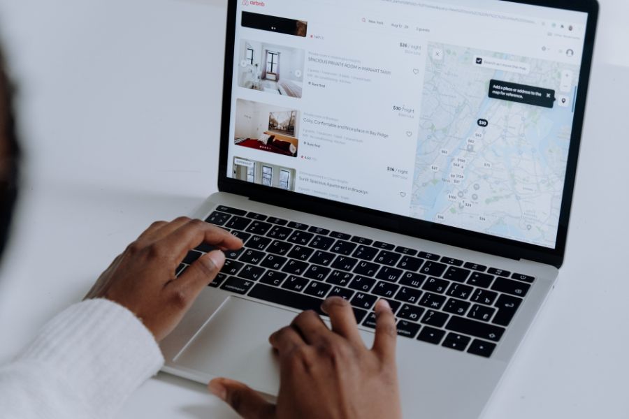 A person browsing Airbnb listings on a laptop, showing a search results page with property photos, prices, and a map of New York City.
