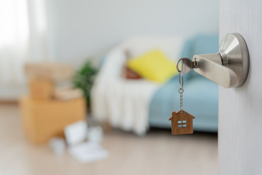A close-up of a silver door handle with a key and wooden house-shaped keyring in the lock, with a softly blurred living room visible in the background.
