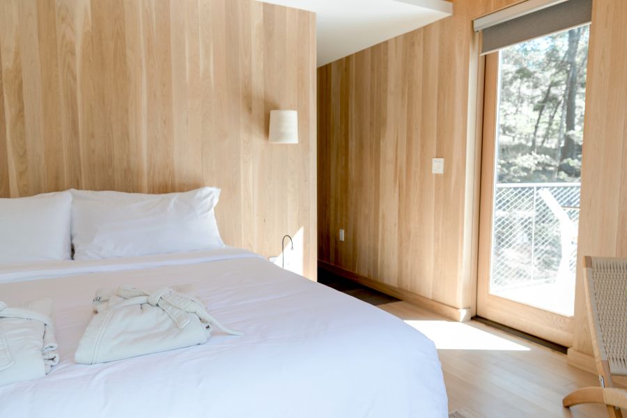 A bright, minimalist hotel room with warm wood-panelled walls, a neatly made white bed, a folded white robe, and a glass door leading to a balcony with trees outside.