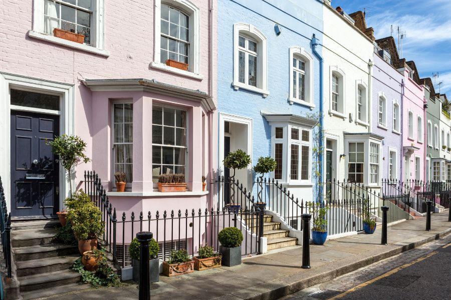 A row of pastel-painted Georgian terraced houses in pink, blue, and lilac along a quiet London street, with black iron railings and potted topiary plants at the front.
