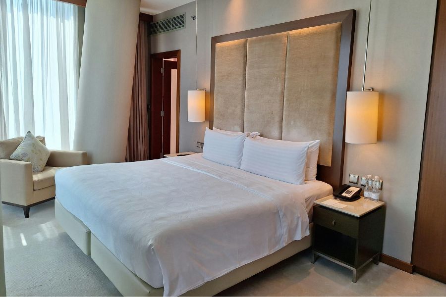A spacious, elegant hotel room with a large king-size bed dressed in crisp white linens, a tall padded headboard, warm bedside lamps, and sheer curtains letting in soft light.