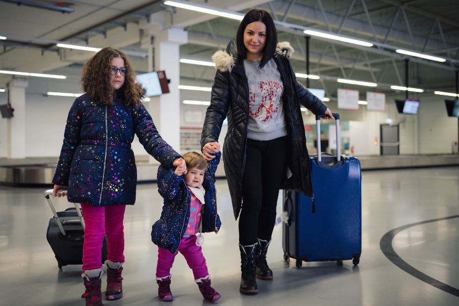 A mother walks through an airport arrivals hall with her two young daughters, pulling suitcases.