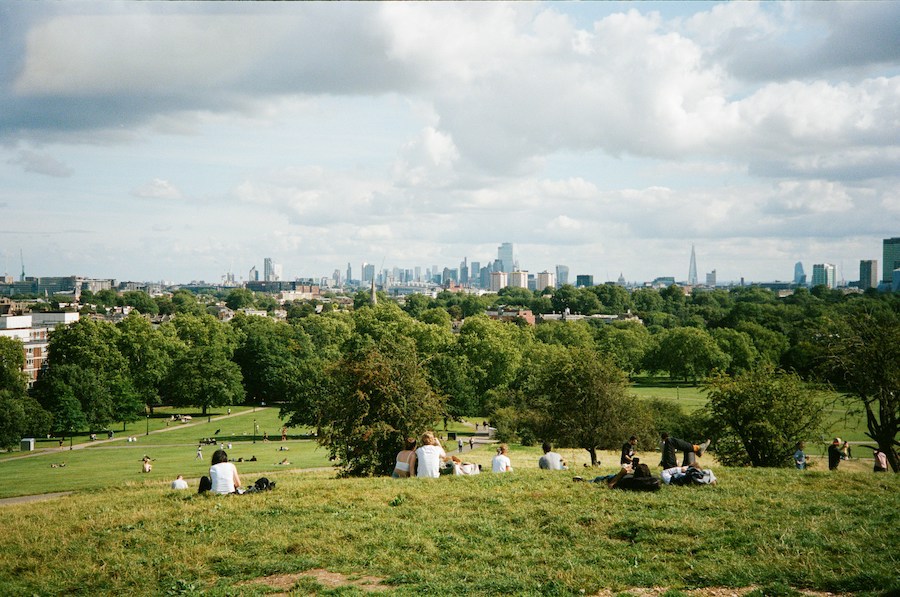 This is an image of Primrose Hill. There are people sitting on the green grass and you can see London's skyline in the distance. It is day time. The London Eye is one of London's most overrated attractions, try Primrose Hill instead.