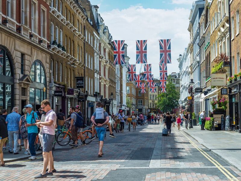 This is a picture of a spacious street in Soho, London. People are milling around the middle and British flags adorn the middle of the street.