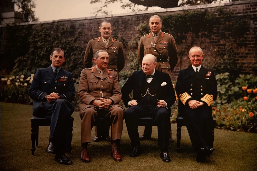 This is an image of Winston Churchill sat taking a group photographer with other men in uniforms in a garden.