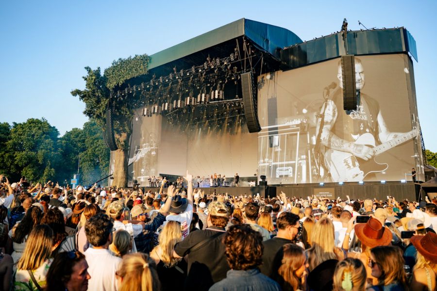 A large outdoor concert at BST Hyde Park on a sunny evening. A massive stage with dramatic lighting rigs and a giant screen displaying a black-and-white image of a guitarist performing. A large crowd fills the foreground, many with arms raised.
