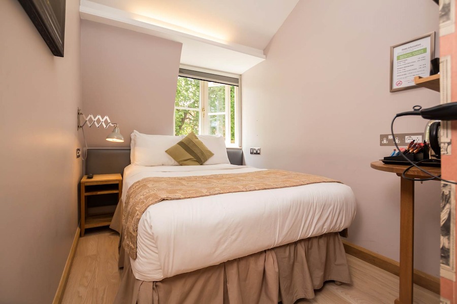 This is a hotel bedroom equipped with a double bed, window, desk and television. The room is toned in muted colours of beige, brown, white and grey and is minimalistic. 