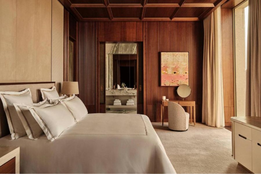 A luxurious hotel bedroom featuring warm wood-panelled walls with vertical fluting, a coffered ceiling, a neatly made king-size bed with ivory bedding, a dressing table with round mirror, abstract pink artwork, and a glimpse into a marble bathroom.
