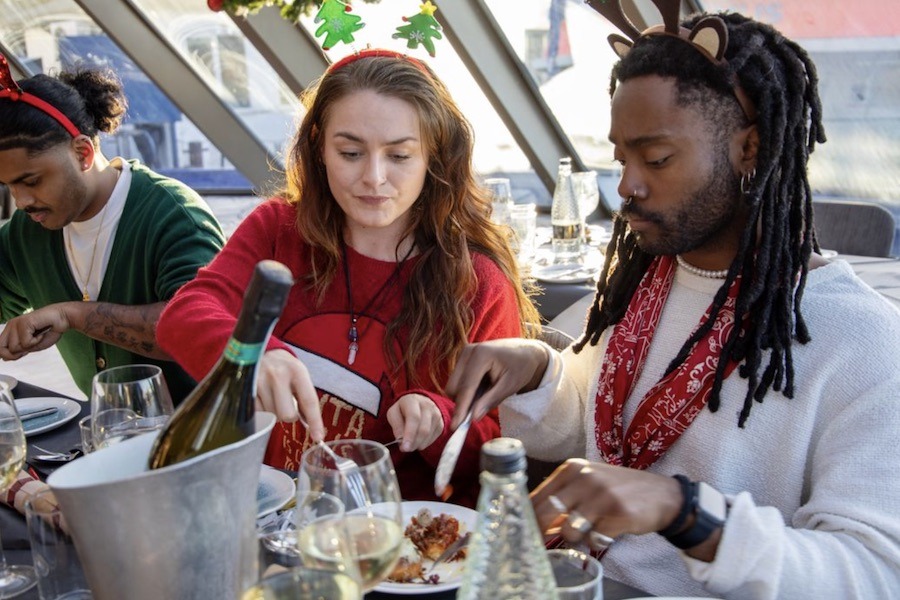 This is an image of people cutting up a turkey and a plate of food on a boat. They all have Christmas hats and jumpers on.