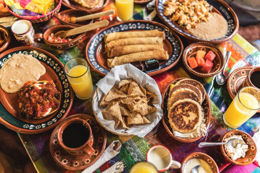 A vibrant spread of traditional Mexican dishes served on hand-painted terracotta pottery, laid out across a colourful woven tablecloth. From crispy taquitos and tortilla chips to refried beans, fresh salsa, fruit, and gorditas.