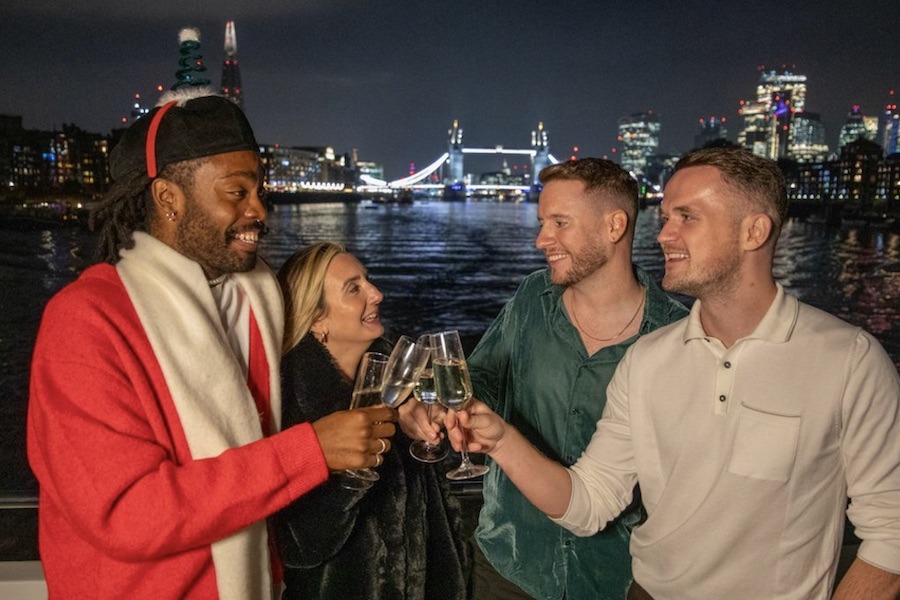 This is an image of people clinking wine glasses on the deck of a boat with a backdrop of a sparkling tower bridge. One of the men is wearing a Santa costume.