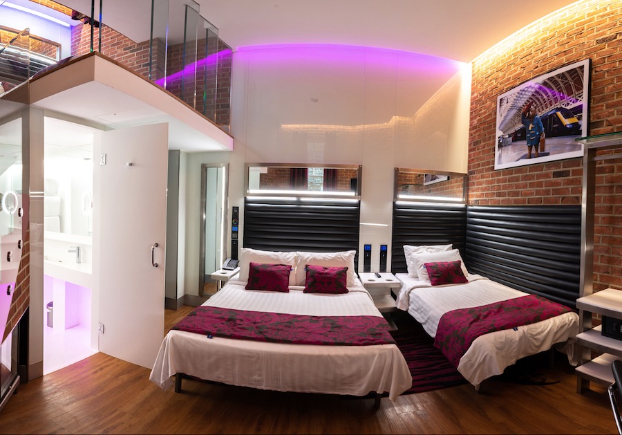 This is an image of a neat hotel bedroom with two beds and cool lighting. The floor is wooden and the walls are white and one wall is made of bricks. There are cool purple lights shining around the room. 