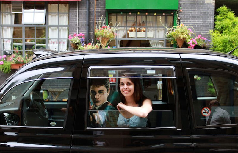 This is an image of a woman sat in a black cab. Next to her is a poster of Harry Potter in his uniform and holding a wand. The woman is smiling and looks very happy.