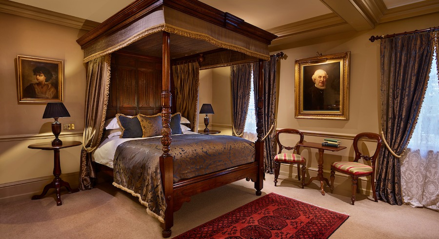 This is an image of a decadent looking hotel bedroom with mahogany furnishings and a big luxurious double bed and carpeted floors with paintings hung on the walls.